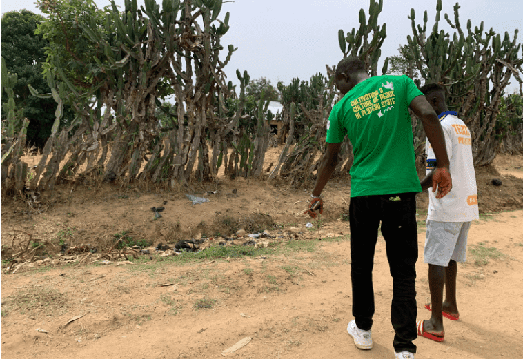 Two people walk along a dirt path beside tall cacti, one in a green shirt and the other in white, outdoors.