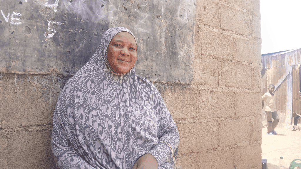 A smiling woman in a patterned hijab leans against a brick wall; a boy stands in the background near metal sheets.