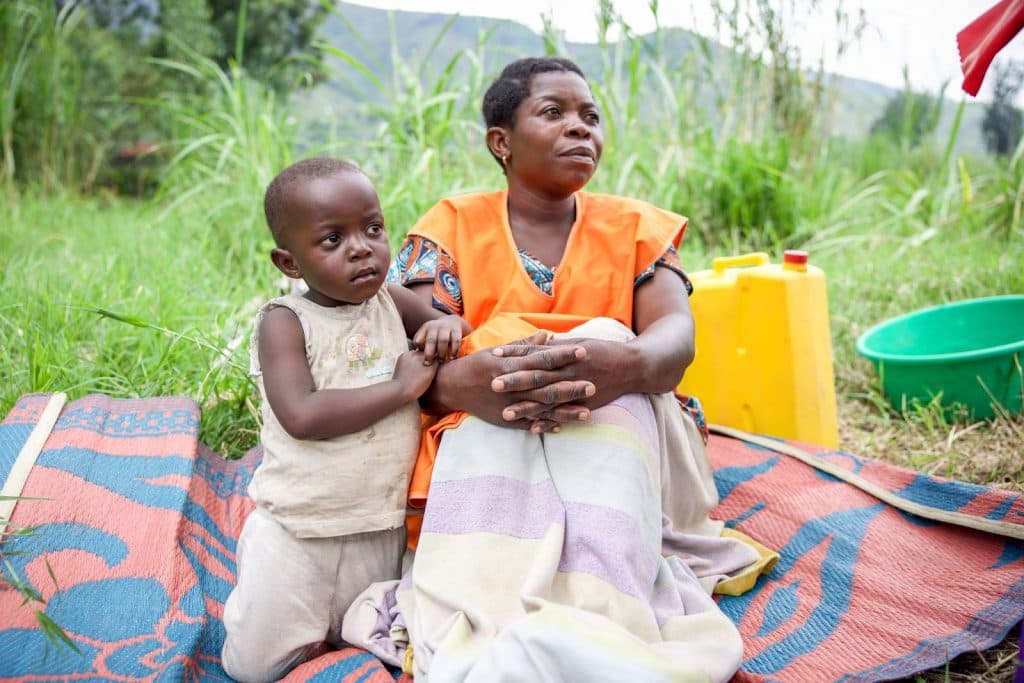 A woman and child sit on a mat in a grassy field, with a yellow container and green basin nearby.