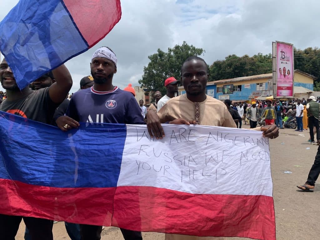 Protesters hold a tricolor flag with a message written on it, amidst a crowd on a cloudy day.