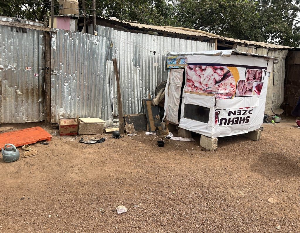 Small makeshift stall with meat images on a dusty ground, surrounded by corrugated metal sheets and various objects.