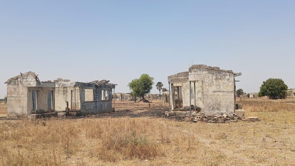 Abandoned, crumbling buildings stand in a dry, grassy field under a clear blue sky, with scattered trees in the background.