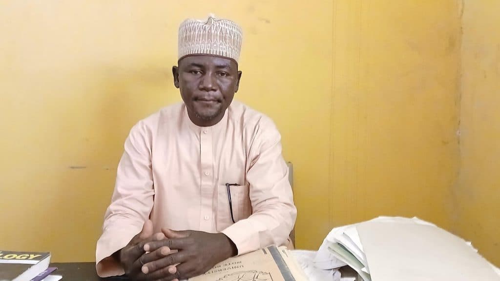 A man in traditional attire sits at a table with books and papers against a plain yellow background.