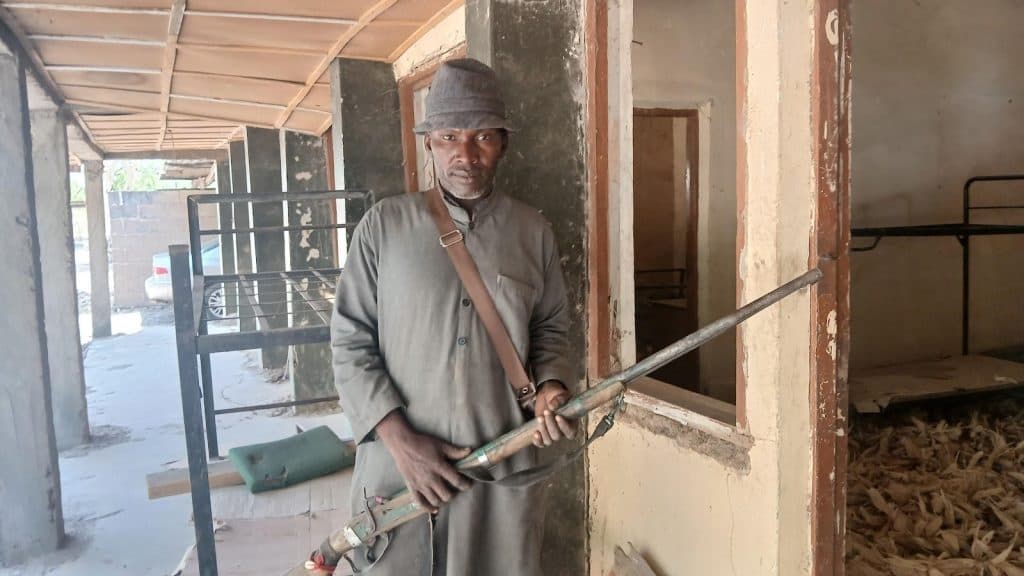 Man in a hat and long robe holding a large pipe stands inside a rustic building with visible wooden and brick walls.