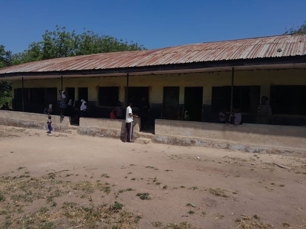 A group of people standing and sitting outside a long, low building with a rusty metal roof on a sunny day.