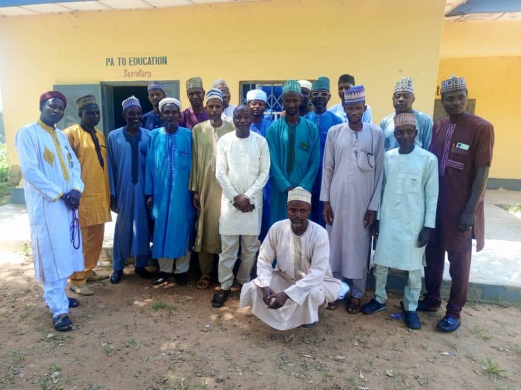 A group of men in traditional attire stands outside a building labeled "PA to Education Secretary."