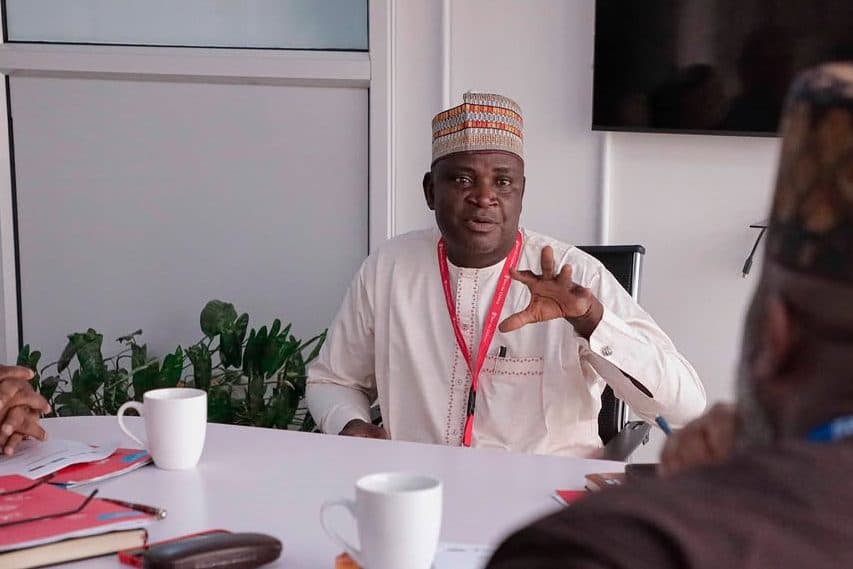 Man in traditional attire gestures during a meeting at a table with two white mugs and documents.