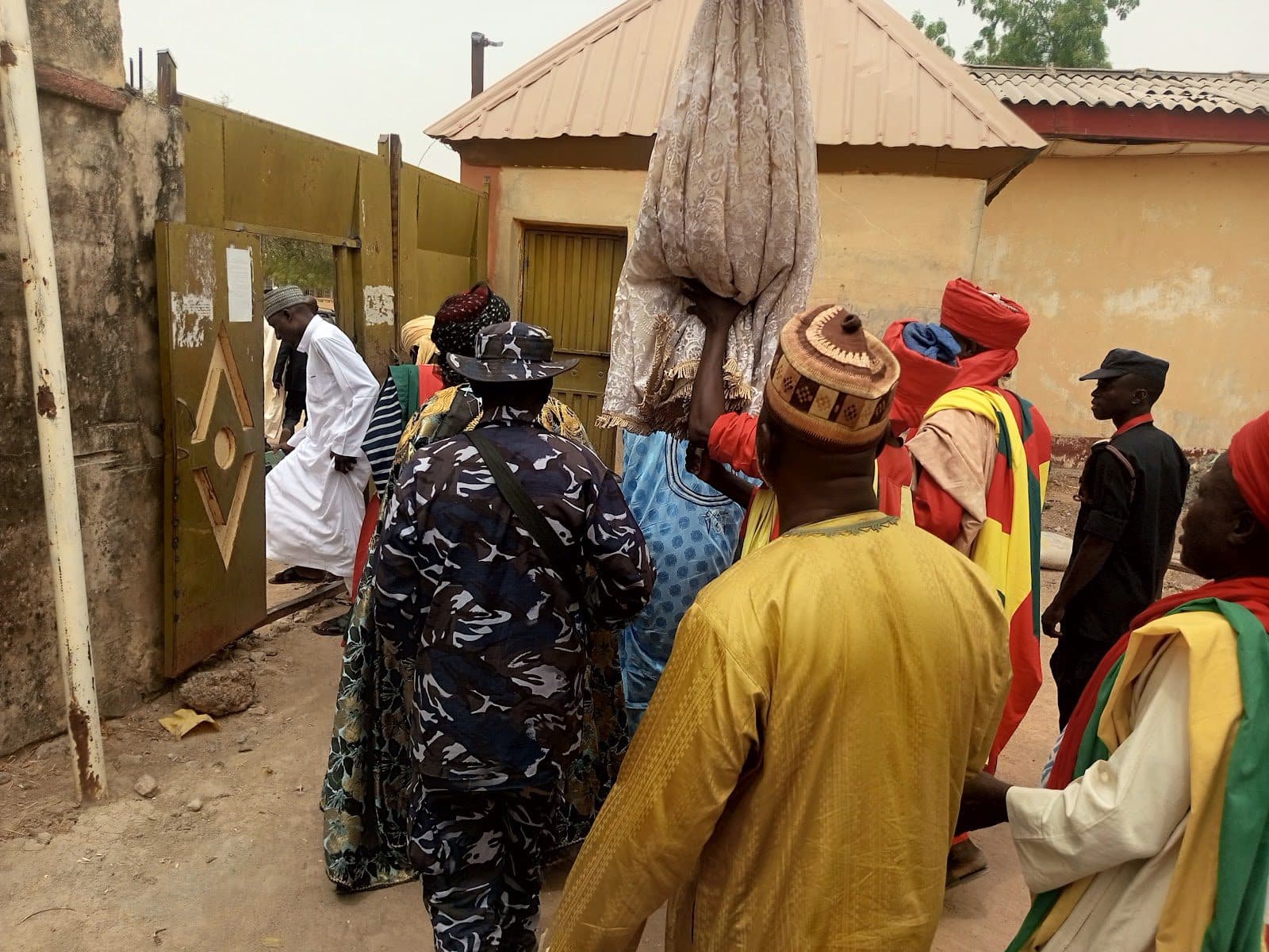 A group of people dressed in colorful traditional attire entering through a large gate, one carrying a tall wrapped object.