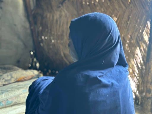 Person in a blue hooded garment sits indoors, facing away. Woven wall texture and soft light create a peaceful atmosphere.