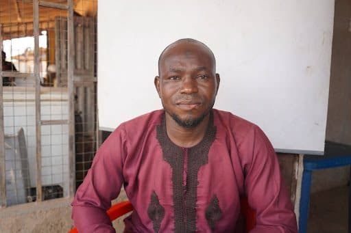 Man in maroon traditional attire sitting indoors, with a subtle smile and a plain background.