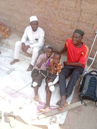 A group of four people sitting on the ground, smiling, with crutches and a backpack nearby, against a brick wall background.