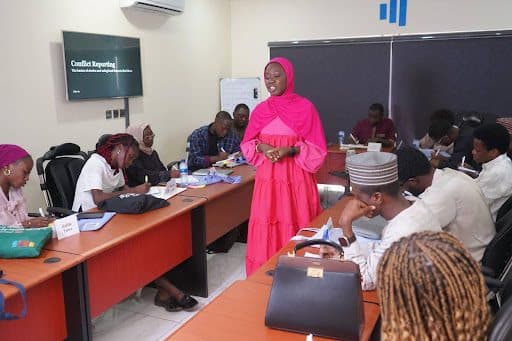 A person in pink speaks to a group seated around tables in a classroom labeled "Conflict Reporting."