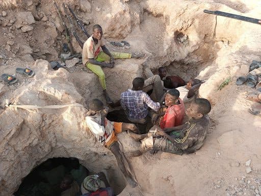 Group of boys sitting in a rocky area with makeshift mining tools around them.