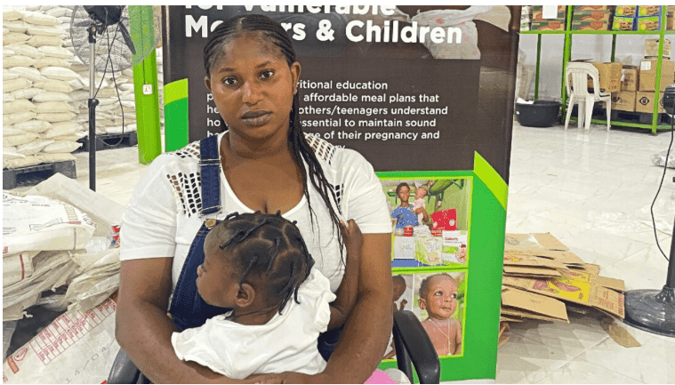 A woman sits with a child in her lap in front of a sign about resources for vulnerable mothers and children, surrounded by bags and boxes.