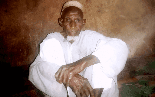Elderly man in white clothes and a cap, seated against a textured wall, with hands resting on his knee.