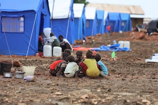 Children sit in a circle on muddy ground near blue tents in a refugee camp.