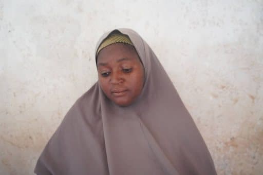 A woman wearing a brown hijab looks down, set against a textured beige wall.