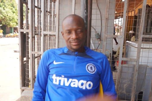 Person wearing a blue Chelsea football jersey sitting outdoors, with a blurred hand in the foreground.