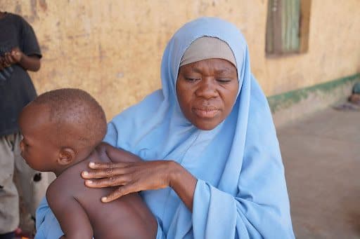 A woman in a blue hijab holds a young child, pressing on the child