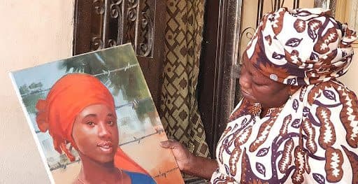 Woman holding a portrait of a young girl in an orange headscarf, both wearing patterned clothing, standing by a doorway.