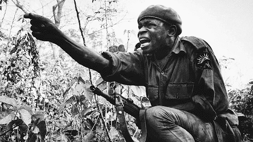 Soldier with a beret, crouched in dense vegetation, points forward with intensity, his rifle slung across his back.