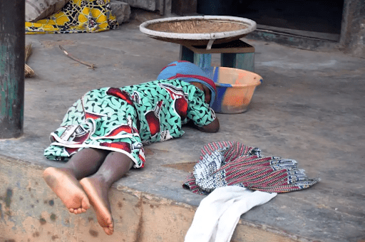 Child in colorful clothing sleeps on a concrete floor, near a plastic bowl and fabric.