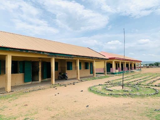 A long building with a corrugated metal roof, open doors, and windows, set in a sandy area with sparse grass and small rocks.