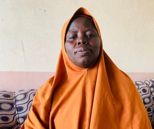 Person in an orange hijab sits on a patterned sofa, facing forward with a neutral expression.