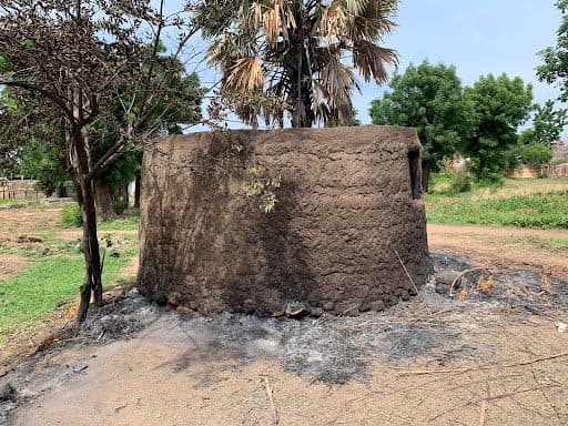 Round mud hut with a thatched roof, surrounded by charred ground and sparse trees in a rural setting.