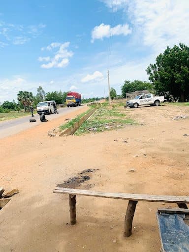 Rural road with parked vehicles, including a truck and a car. Dirt foreground, trees lining the road, and a partly cloudy sky above.