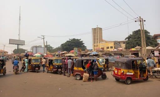 Busy street scene with multiple parked rickshaws, vendors, and people walking around under a clear sky. Buildings and power lines in the background.