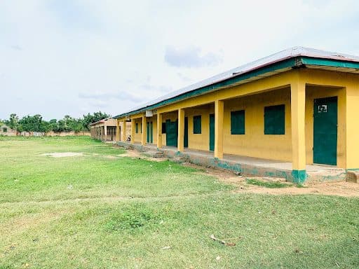 Long, yellow school building with green doors and windows, set on a grassy field under a partly cloudy sky.