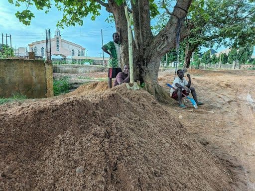 Three people resting by a tree near a dirt pile, with a church visible in the background.