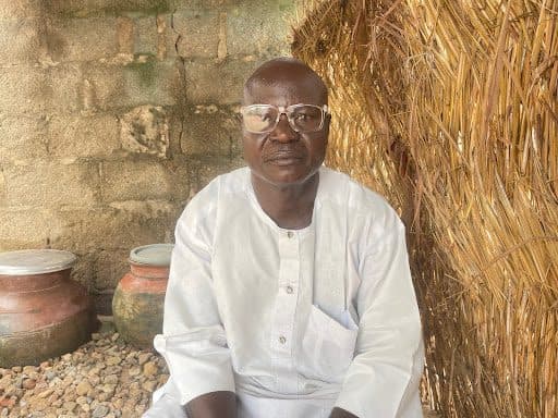 Man in white attire, wearing glasses, sitting on gravel with pots and a straw wall background.