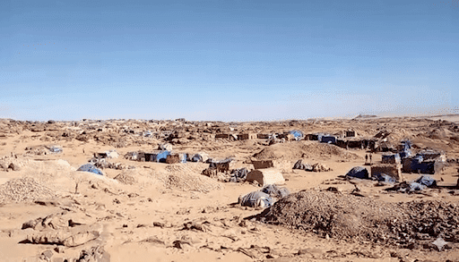 Desert landscape with makeshift tents and structures scattered across sandy terrain under a clear blue sky.