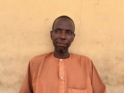 Man in an orange shirt standing against a textured beige wall, looking slightly to the side.
