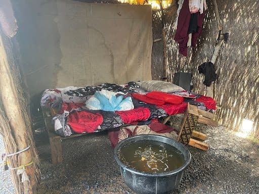 A makeshift bed with colorful blankets inside a rustic shelter, and a tub of water on the gravel floor nearby.