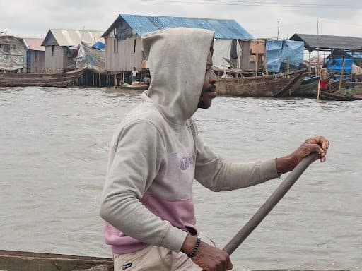 Person in a hoodie paddling a boat on a river, with stilt houses in the background.