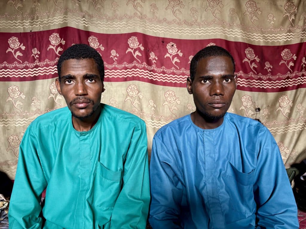 Two men in colorful shirts sit against a floral patterned curtain backdrop, looking directly at the camera.