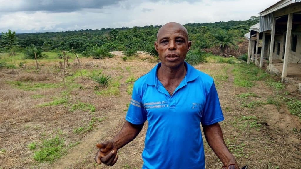 Man in a blue shirt standing by a field and building, with lush trees in the background under a cloudy sky.