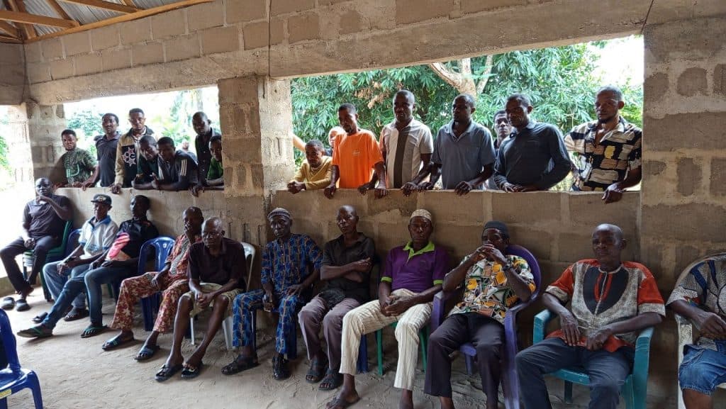 A group of men sit and stand inside a partially constructed brick building with a tin roof, some looking at the camera.