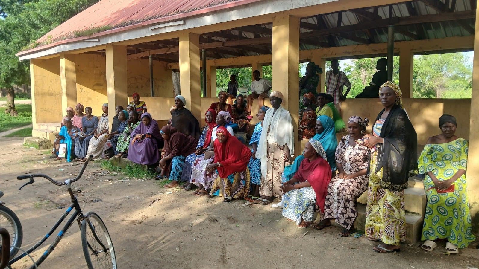 A group of people, mostly women, sit and stand outside a building with a rusty roof. A bicycle is parked in the foreground.