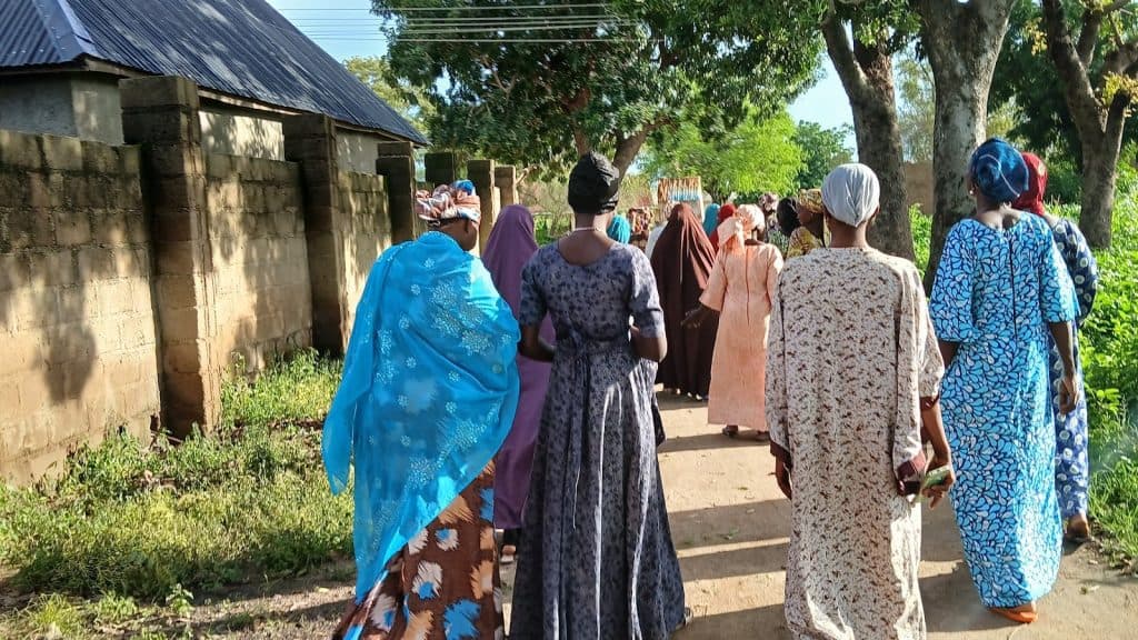 A group of colorfully dressed people walk along a concrete path bordered by trees and a stone wall, under a sunny sky.