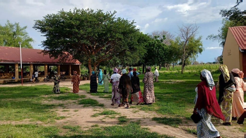 A group of people in colorful attire walk towards a building with a red roof, surrounded by greenery and trees under a cloudy sky.