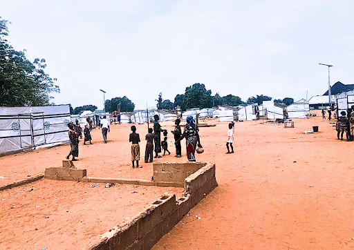 People gather in an open area of a camp with tents and structures in the background under a cloudy sky.