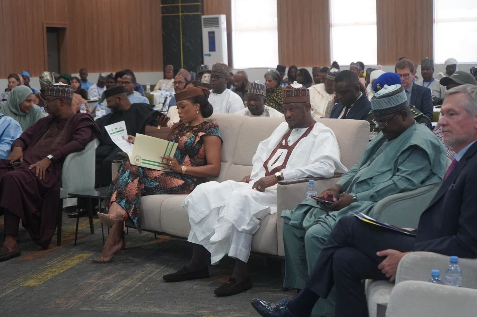 People seated in a conference room, engaged in reading and discussions, with documents and water bottles on tables in front of them.