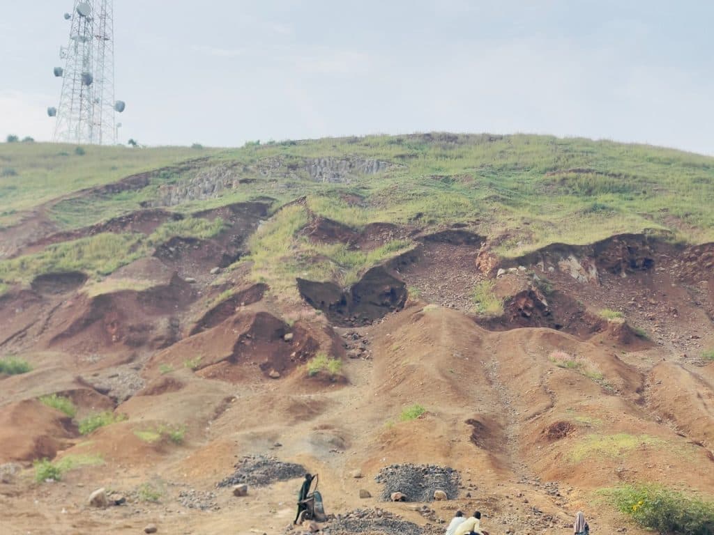 Hilly landscape with patches of green, rocky terrain, and people seated below a communication tower; overcast sky above.