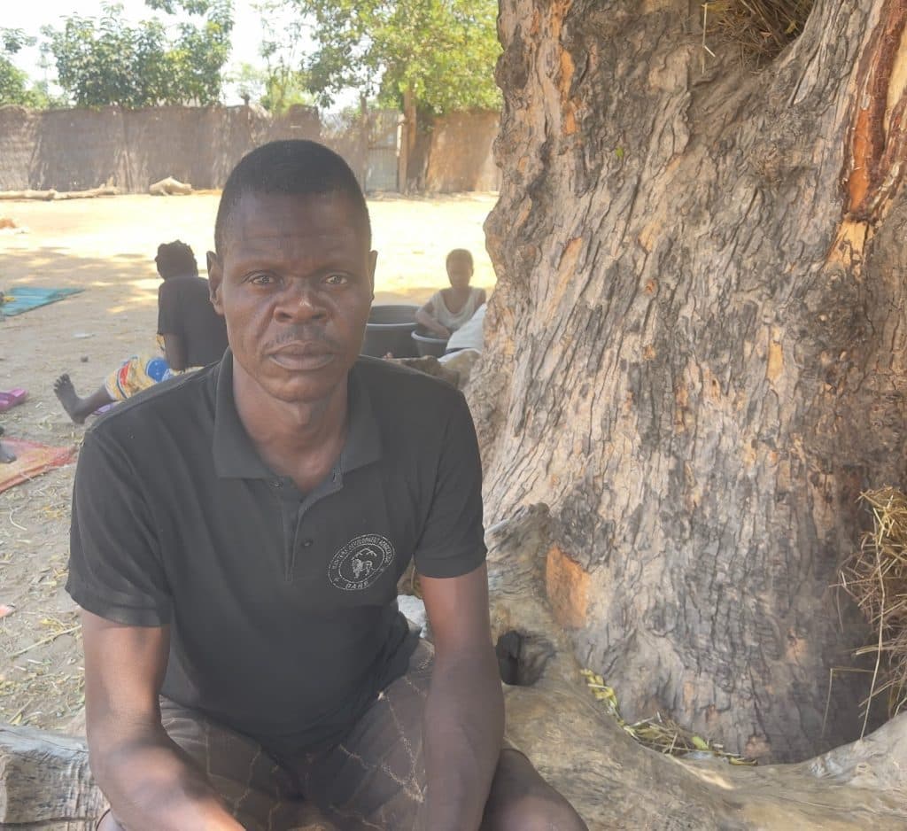 A man in a black shirt sits by a large tree with people and woven mats in the background, set outdoors on a sunny day.