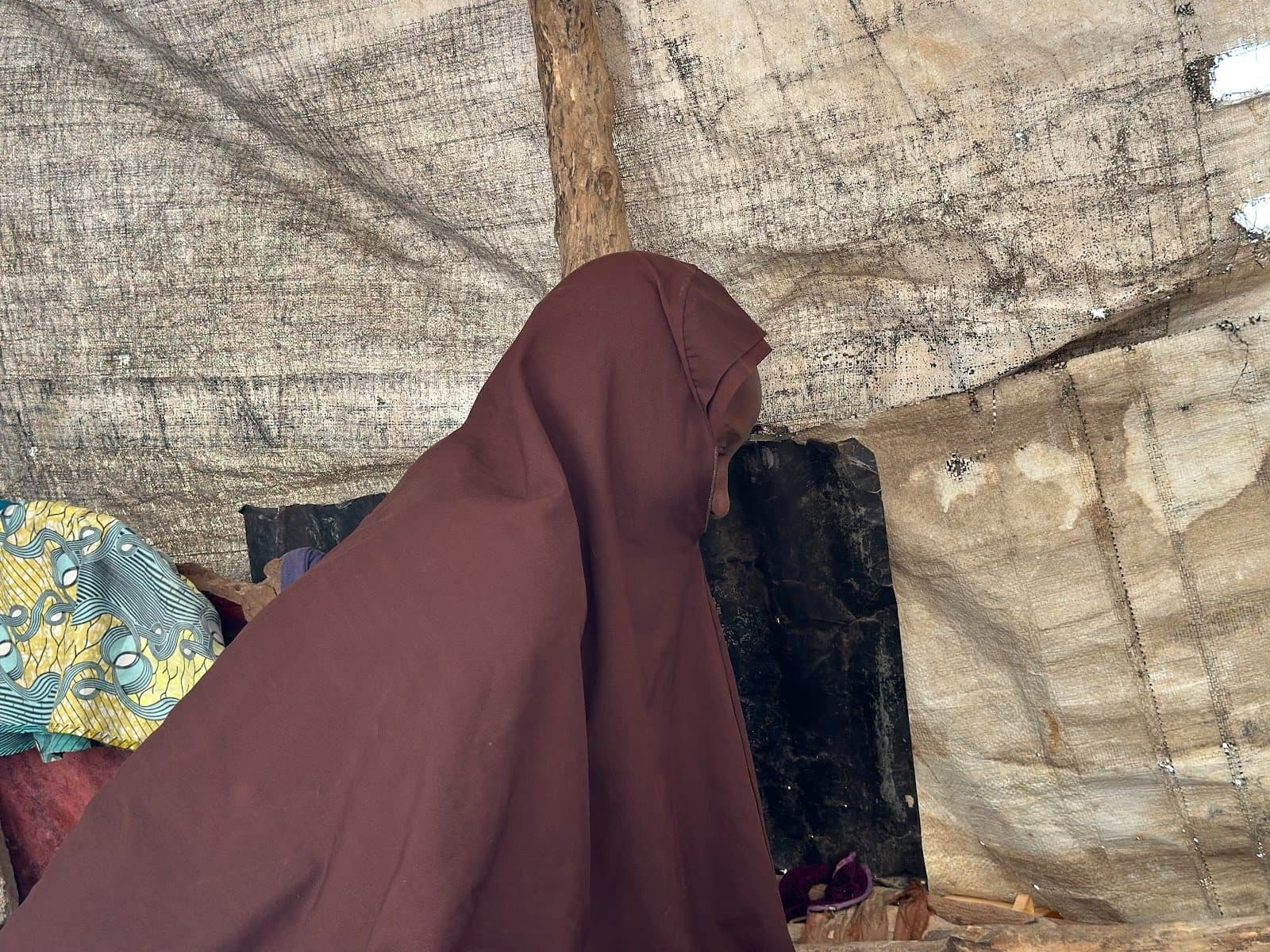 Person in a brown cloak sitting inside a rustic, tarp-covered shelter.