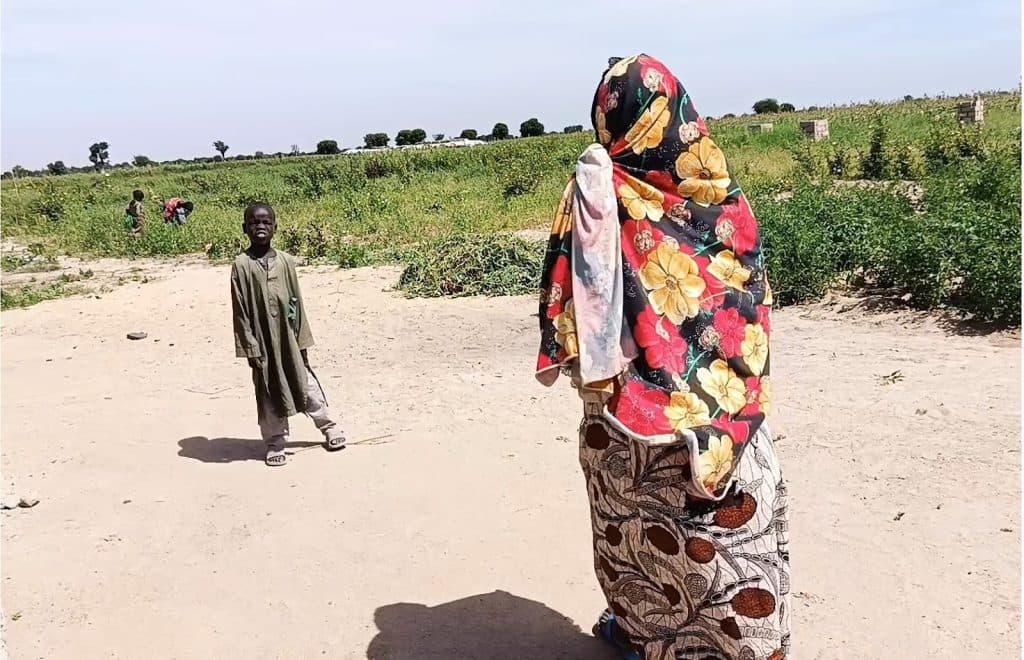 A child and a person in a floral shawl stand on a dusty path near a field, with others and green plants in the background.